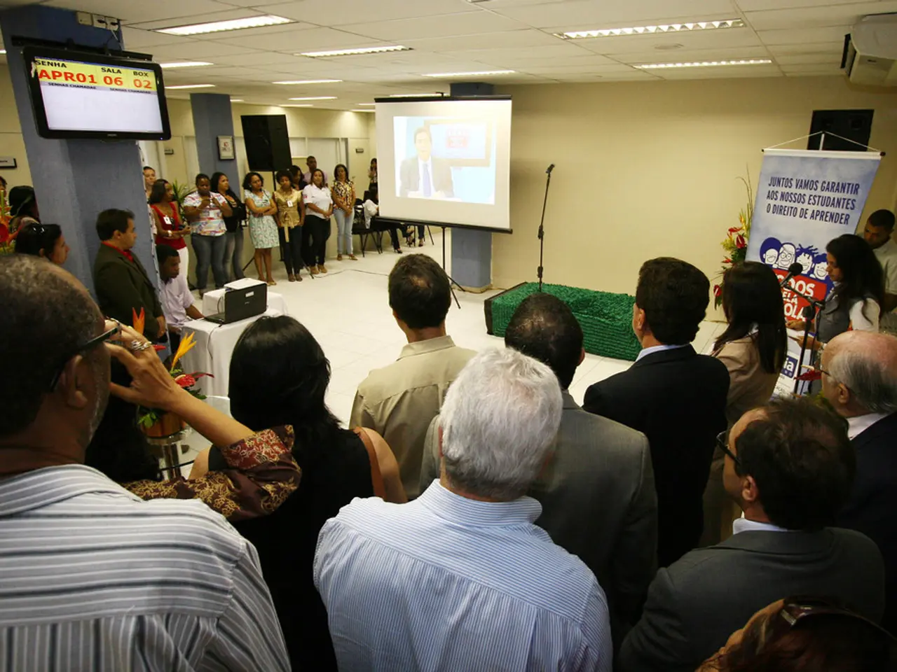 In the image we can see there are lot of people standing and there is a projector screen in the...