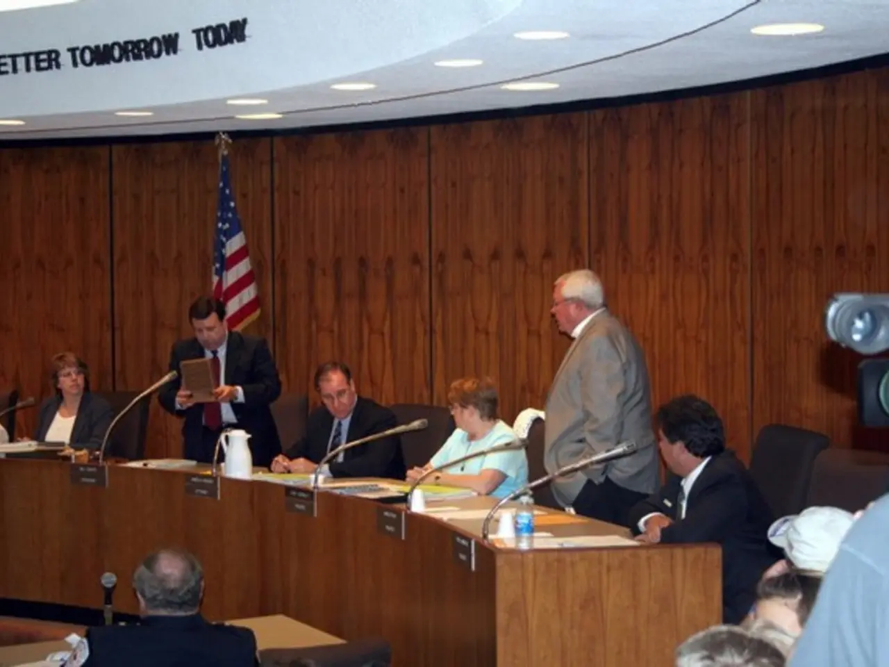 In this image there are group of people sitting on chairs in front of table, behind them there is a...