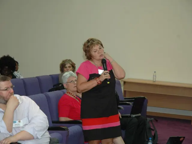 The picture is taken in a room. In the foreground of the picture there are people, chairs, desk,...