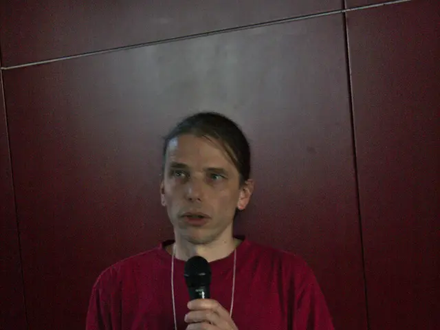 In this picture a red shirt guy is holding a mic in his hand and talking. To the left side of the...