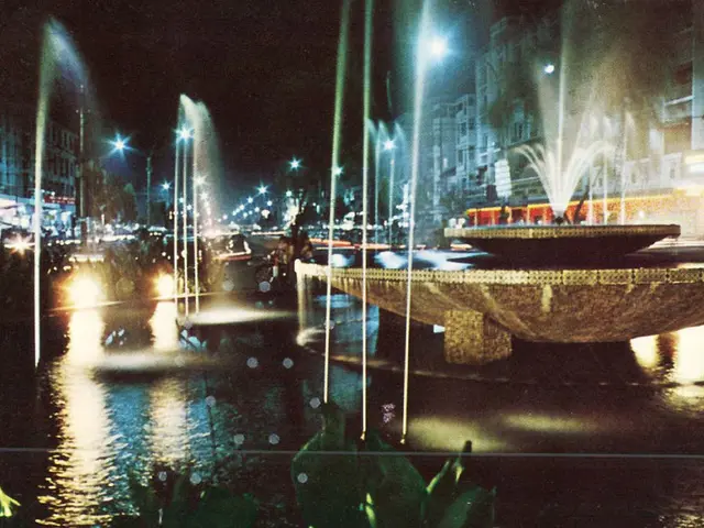 In this image we can see buildings with windows, poles, lights and water fountain.